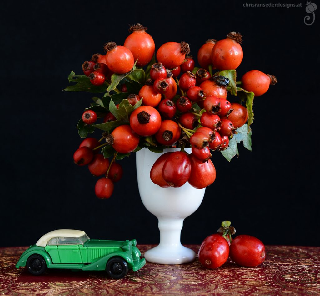 Red berries in a white goblet and tiny model of a green car, placed on an red bookcover. | Rote Beeren in einem weißen Pokal und ein Modellauto stehen auf einem roten Buchcover.