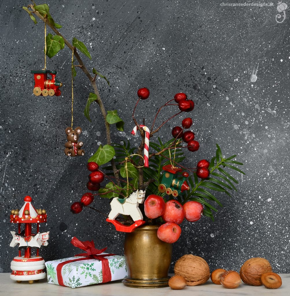 A metal vase with twigs bearing wooden Christmas ornaments bracketed by nuts and a festively wrapped parcel. | Zweiglein mit hölzernem Weihnachtsschmuck füllen eine Vase aus Messing. Rechts von dieser liegen Nüsse, links ein in festliches Papier gehülltes Paket. 