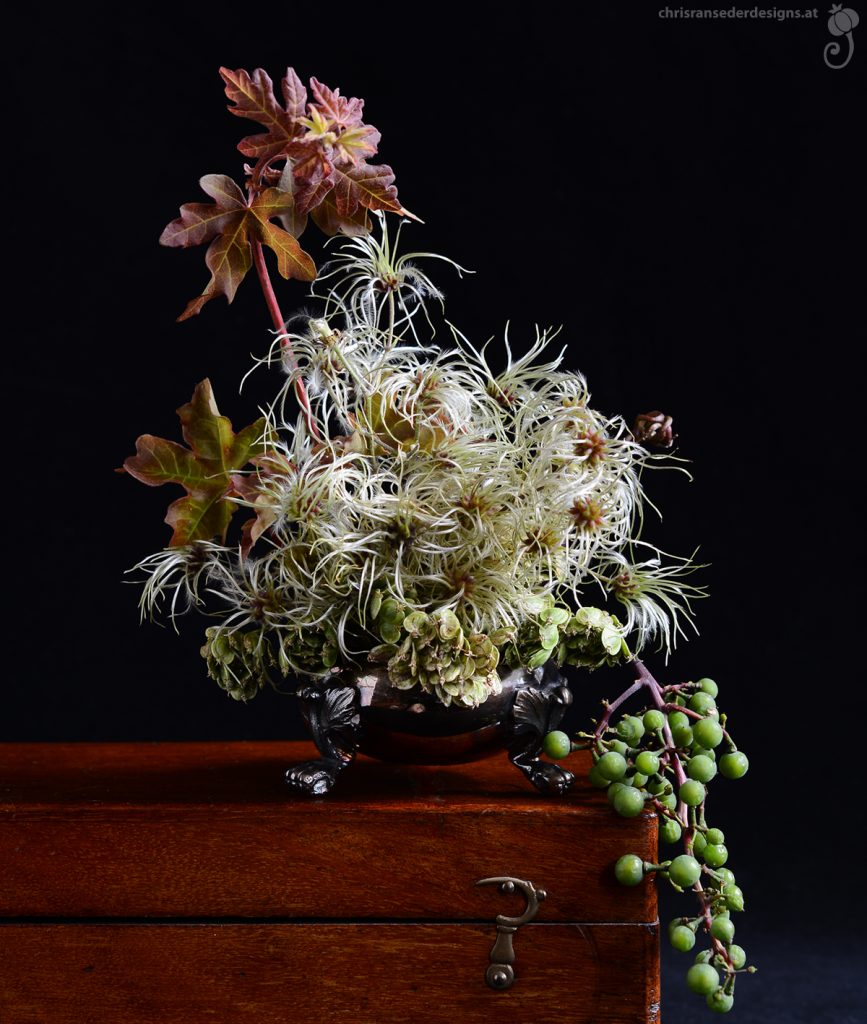 Autumnal seed heads and leaves presented in an saltcellar with clawed feet sitting on a wooden box. | Herbstliche Fruchtstände und Blätter in einem Salzgefäß mit Klauenfüßen, das auf einer Holzschachtel steht.