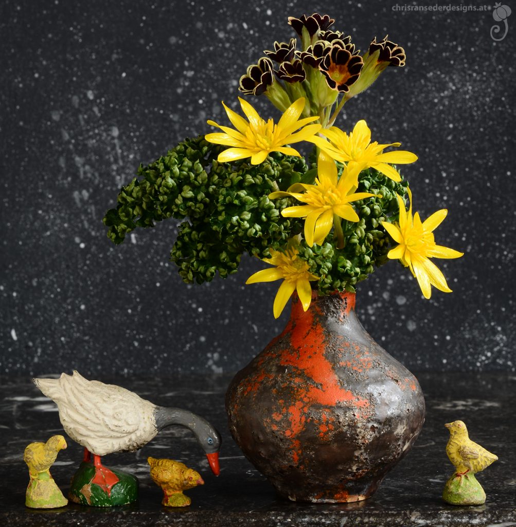 Green vase with yellow flowers and green foliage flanked by vintage toys - a goos and three chicks. | Grüne Vase mit gelben Blumen und grünem Laub, flankiert von altem Spielzeug - einer Gans und drei Kücken. 