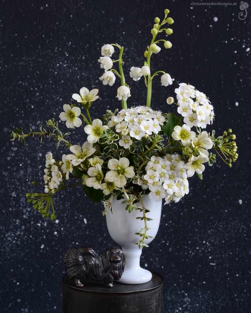 A white goblet filled with white flowers is guarded by a tiny dog made of metal.| Ein weißer Pokal, gefüllt mit weißen Blumen. Davor steht eine winzige Hundeskulptur aus Metall.