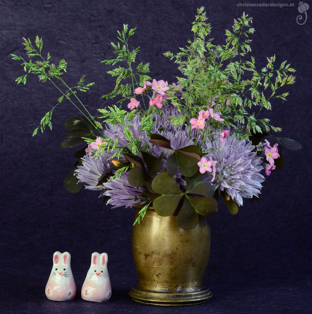 Two ceramic bunnies sheltering under a bunch of purple and pink flowers.| Zwei Häschen aus Keramik unter einem Strauß aus lila und rosa Blumen.