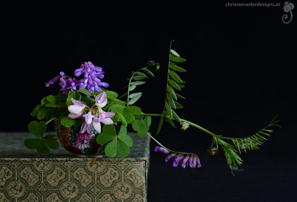 A tiny chinese cup with just two sprigs of flowers and clover leaves standing on a chinese box. | Chinesisches Schälchen gefüllt mit zwei Sorten Blumen, das auf einer chinesischen Schachtel steht.
