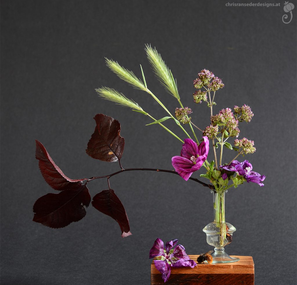 A tiny glass vase filled with flowers and leaves in shades of purple and dark red. | Kleine Glasvase  gefüllt mit Blumen und Blättern in Rot- und Violetttönen.