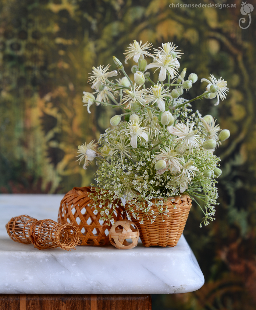 A miniatur basket holds white flowers. Next to it stand more baskets and a tiny fish trap. | In einem kleinen Korb steht ein Strauß weißer Wildblumen, daneben befinden sich weitere Miniaturkörbe und eine Fischreuse. 