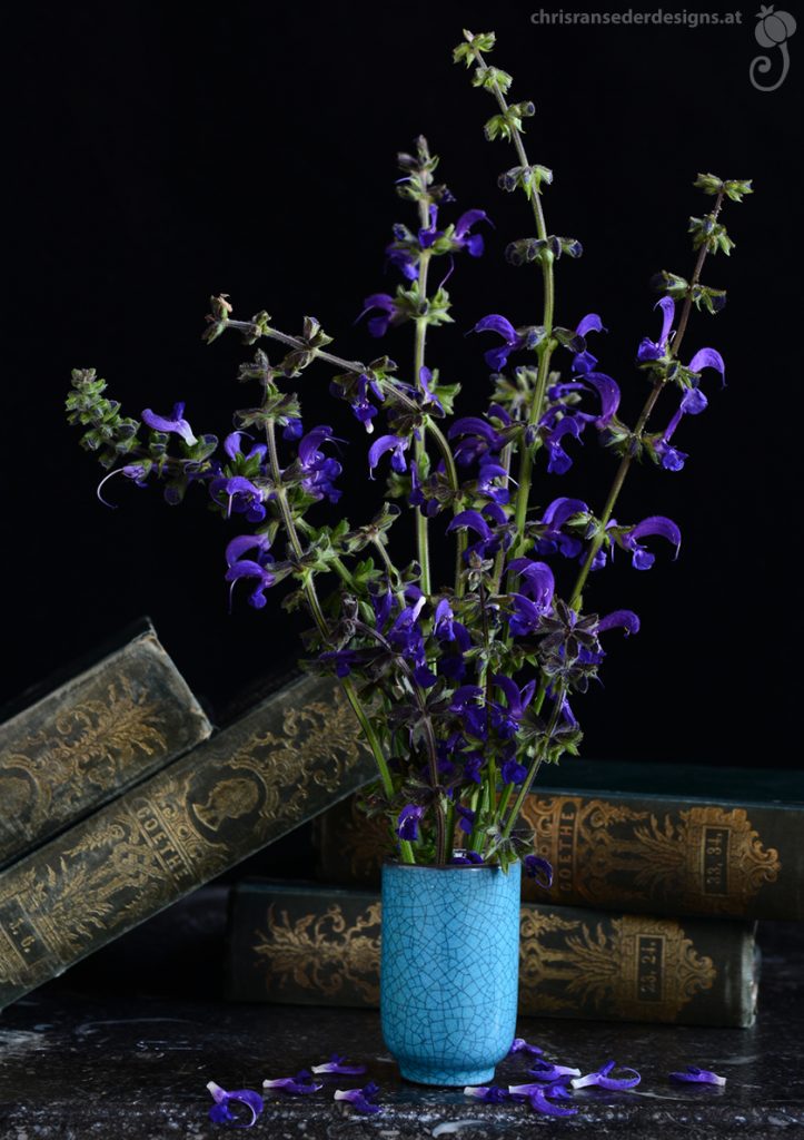 Blauer Wiesensalbei in einer türkisen Vase. Hinter dem Blumenstrauß stehen grün gebundene Bücher.