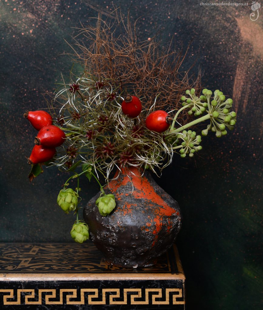 A Bouquet in a spherical vase glazed in grey and orange stands on a patterned wooden box. | Ein in Rot- und Grüntönen gehaltenes Bouquet in einer grau und orange glasierten, kugeligen Vase steht auf einem gemusterten Holzkistchen vor grünem Hintergrund.