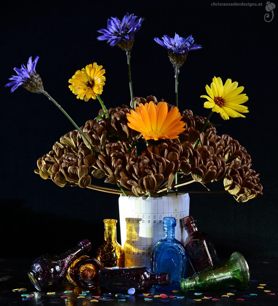 A tiny bouquet of yellow and blue flowers jutting out of a dry seedhead shaped like an  umbrella. At the foot of the vase, wrapped in a page of  an old calender, empty bottles glint in shades of yellow, blue and purple.
