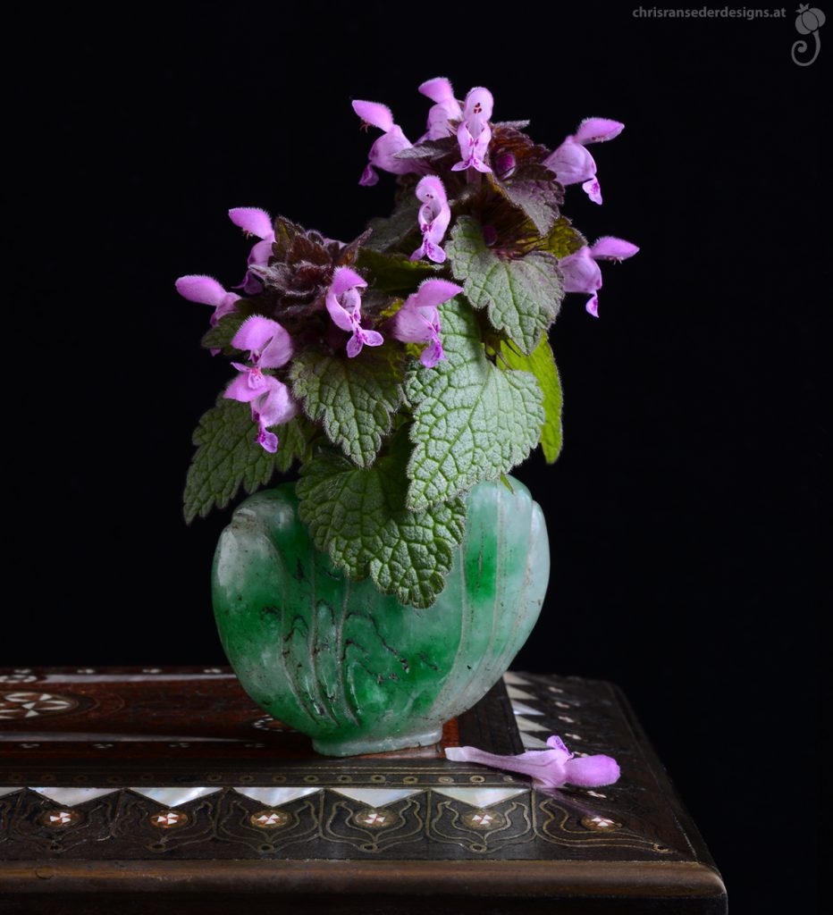 Purpurfarbener Lippenblütler in einem Parfumfläschchen aus grünem Stein. | Purple flowers in a perfume bottle made of green stone.