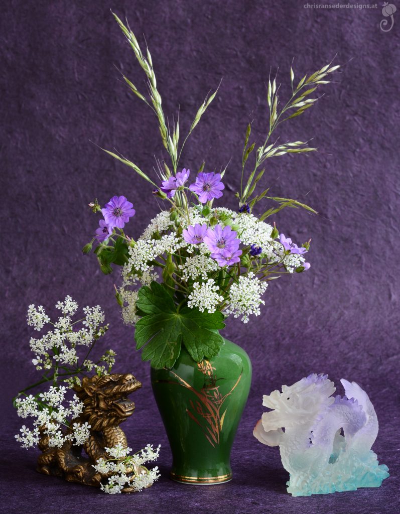 Eine bauchige, grüne Vase gefüllt mit weißen und violetten Blumen steht vor einem lila Hintergrund. Links der Vase versteckt sich ein bronzener Drache in weißen Blütendolden, rechts hält ein violetter Drache die Glücksperle hoch.