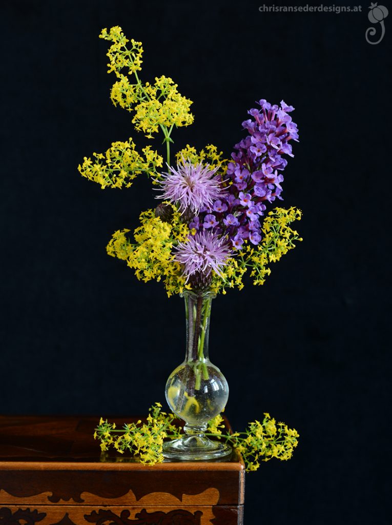 In einer kleinen Glasvase mit kugeligem Bauch stehen gelbe und violette Blumen. der Blumenstrauß steht vor schwarzem Hintergrund auf einem Holzkistchen.