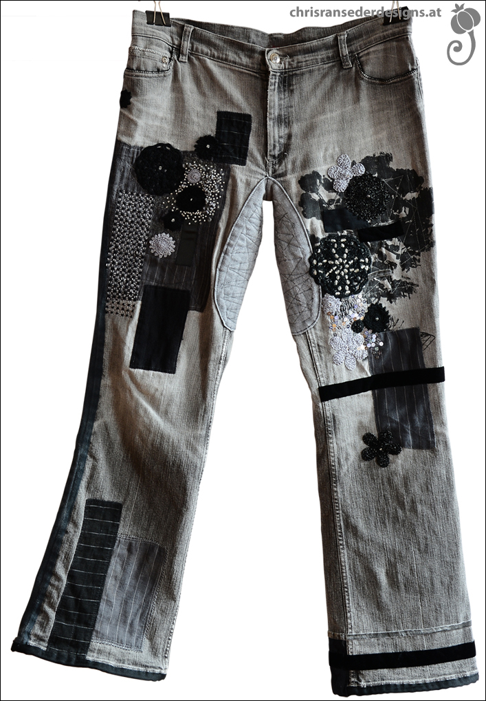 Eine graue Jeans, kunstvoll mit grauen Stoffresten sowie schwarzen und hellgrauen Häkelblumen geflickt.