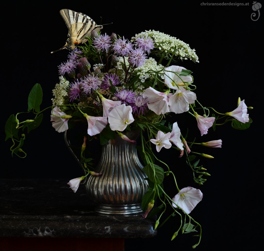 Ein wildes Sträußchen aus weißen und violetten Blumen vom Wegesrand in einem silberfarbenen Kännchen. Ein Schmetterling nascht an den Blüten.