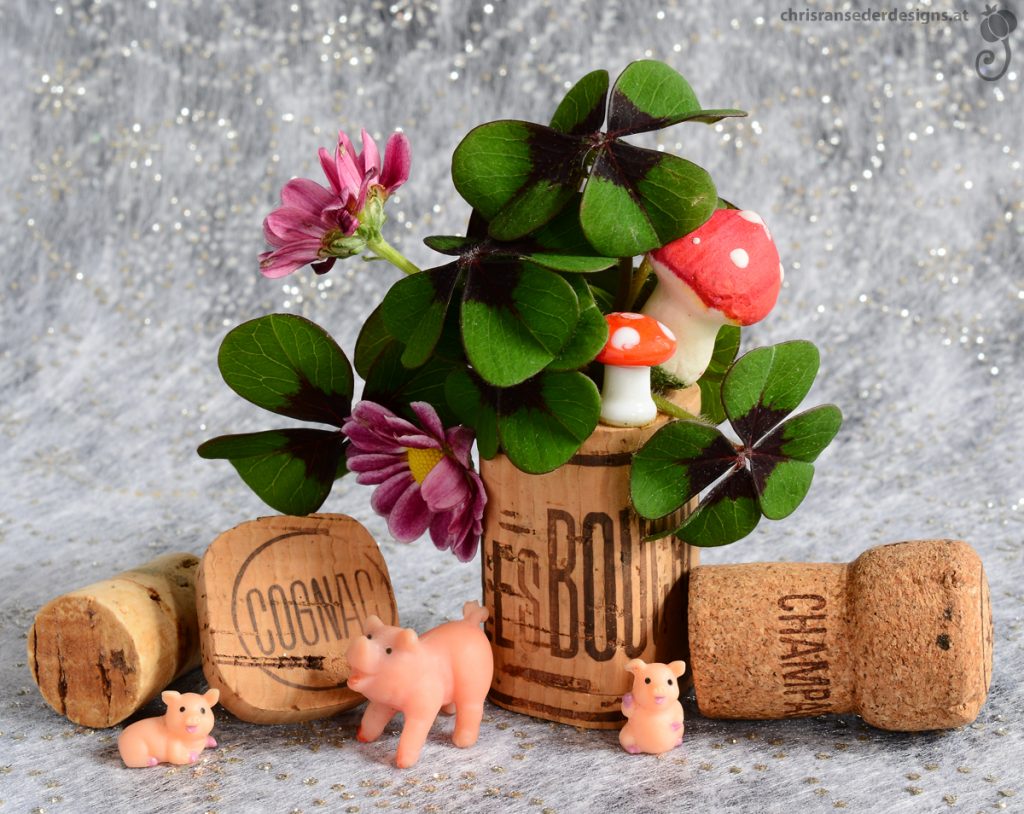 A bunch of clover and pink flowers in a vase made from a bottle cork. Two toadstools sit on the rim. Pigs are foraging next to more corks.