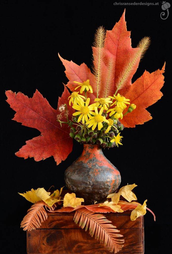 Rot und gelb verfärbtes Laub sowie gelbe Blumen in einer kugeligen, orangerot und grau glasierten Vase.