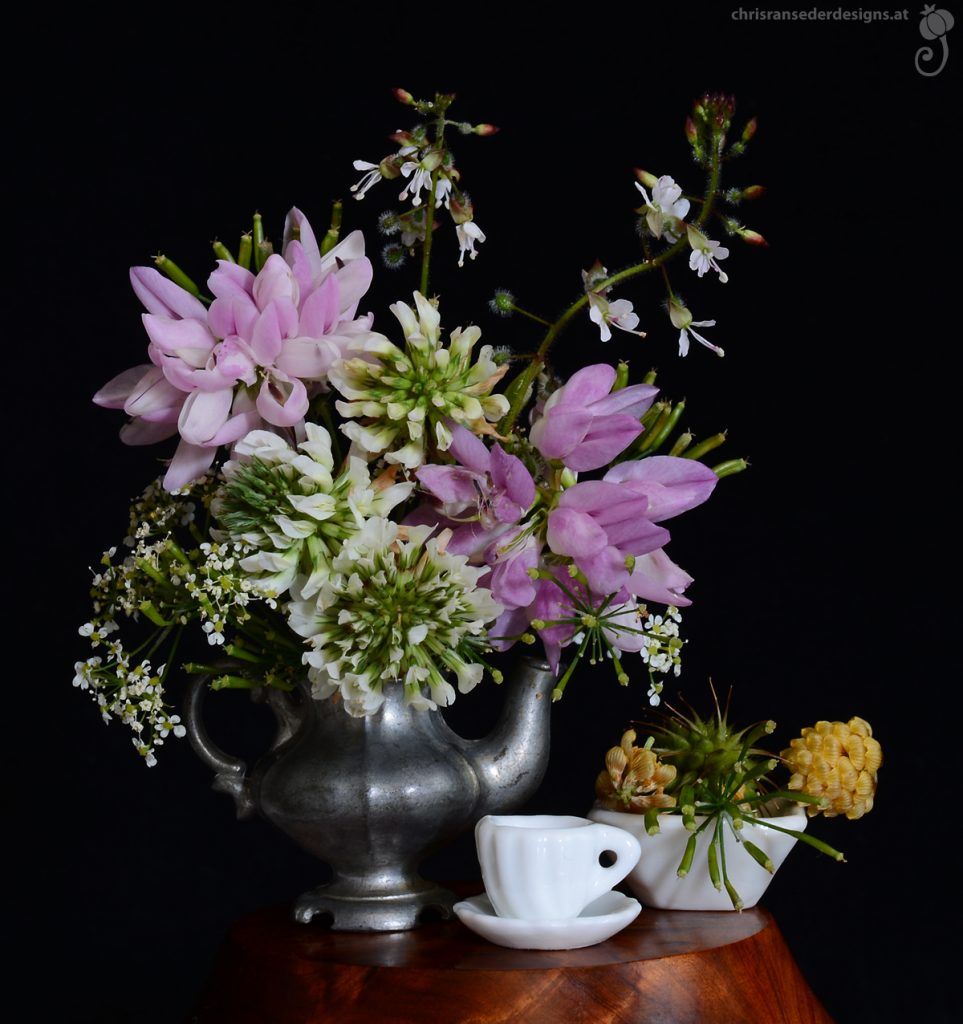 Weiße und hellrosa Wiesenblumen in einer Teekanne aus Zinn.  Daneben weiteres Puppengeschirr: eine Tasse mit Untertasse und eine viereckige Schüssel mit gelben und grünen Samen von Wildblumen.