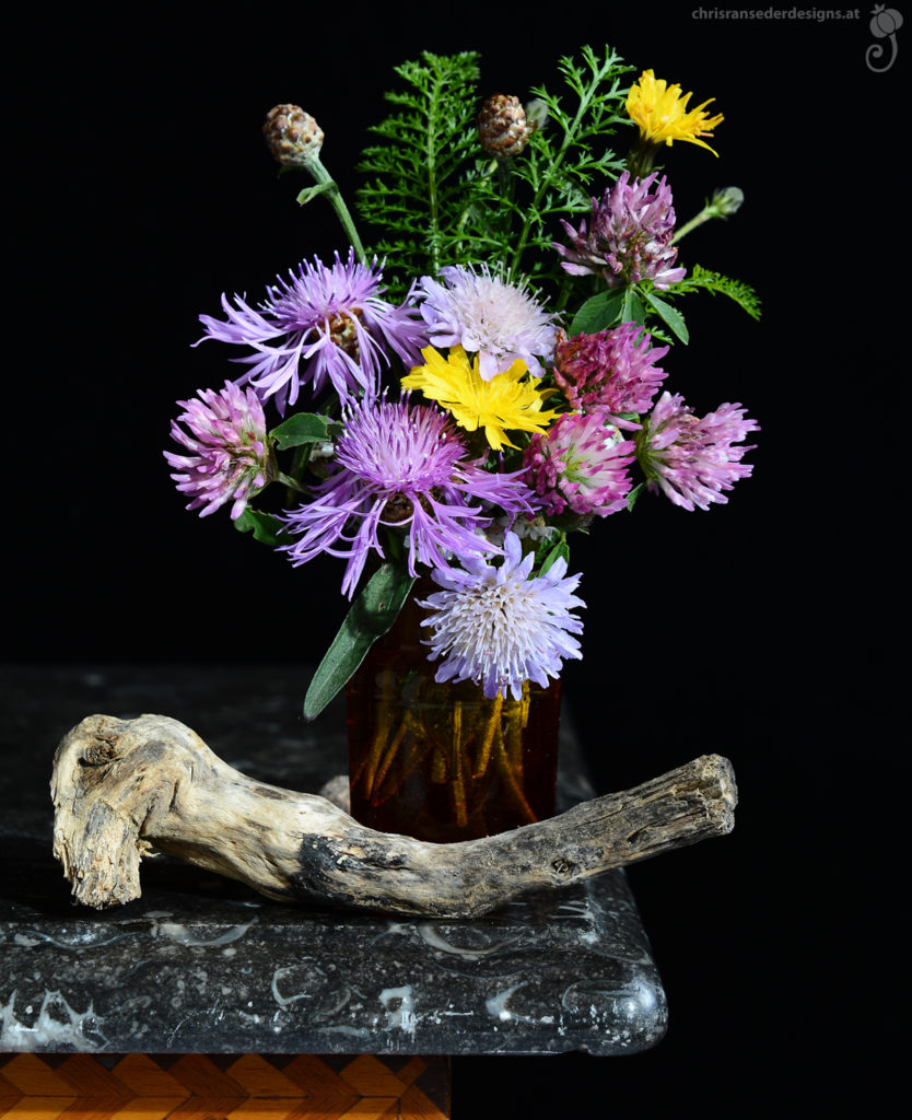 Rote, violette und gelbe Wiesenblumen in einem braunen Medizinglas. Davor liegt ein krummes Aststück ohne Rinde auf der schwarzen Marmorplatte eines Schränkchens.